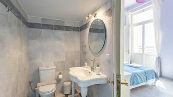 A bathroom with a toilet, sink, and mirror. There is a bed with a blue bedspread in the adjacent room.