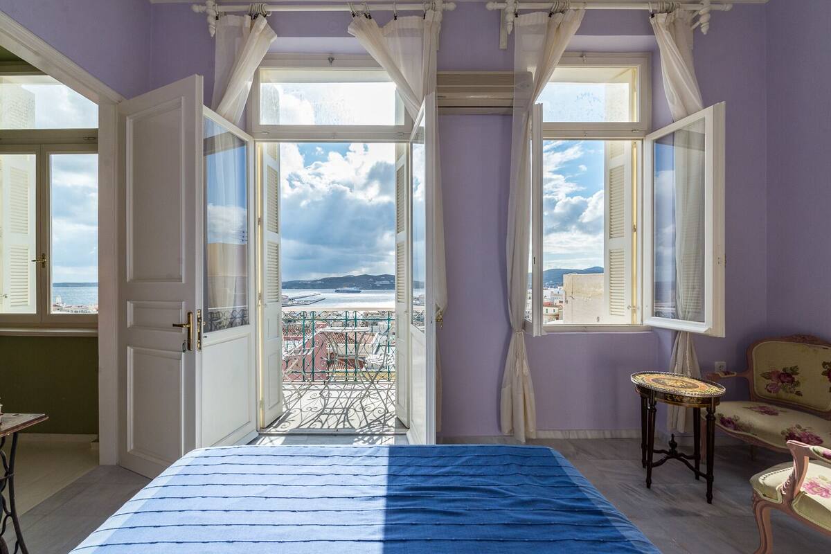 A room with a bed, a chair, and a view of the sea through open windows.