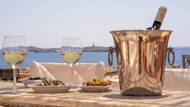 A table set with two glasses of white wine, a silver pot, and plates of food, overlooking a coastal view with a distant island. - Villa Maria (Syros)