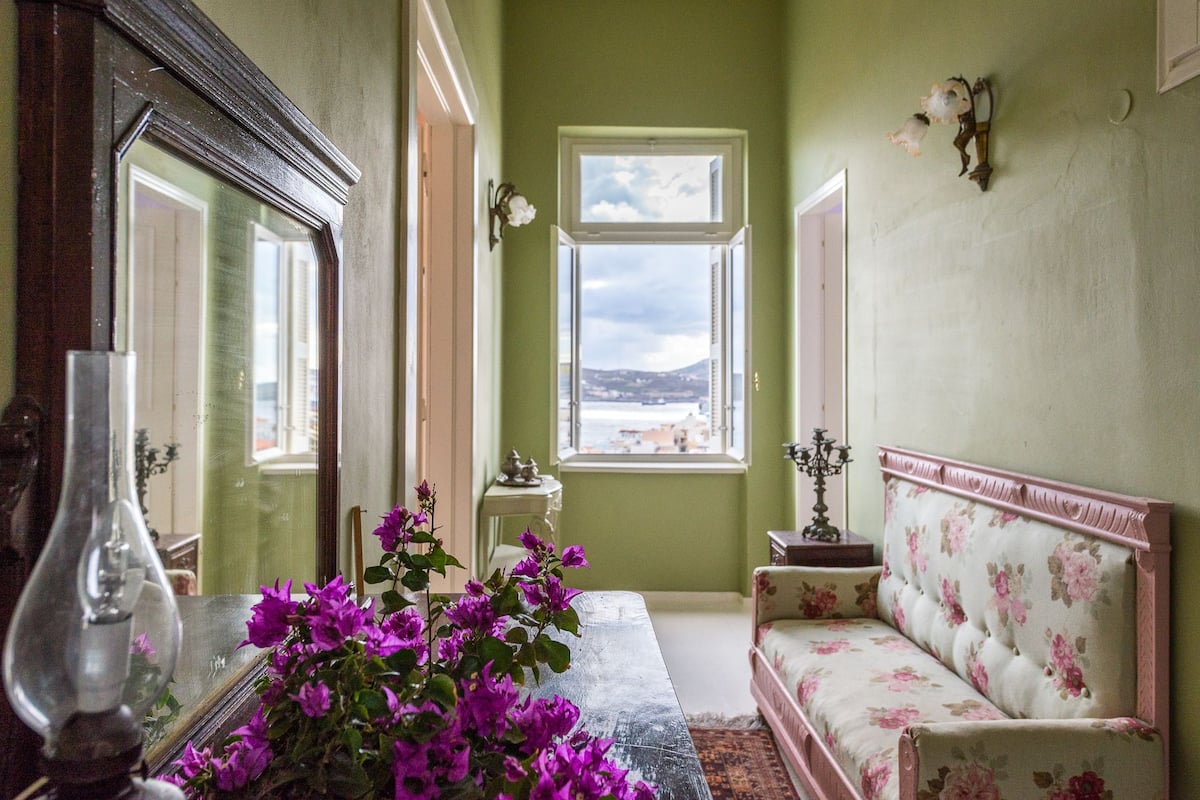 A room with a large mirror, a vase with purple flowers, a sofa with floral upholstery, and a view of the sea through a window.