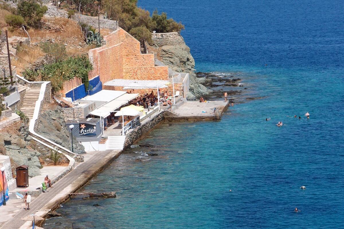 A coastal pier with clear blue waters, a stone building with a sign, and people engaging in various activities.