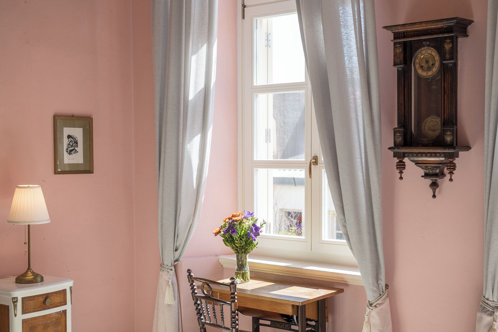 Une pièce avec une fenêtre, une table ornée d’un vase de fleurs, une lampe, une chaise et une horloge fixée au mur.