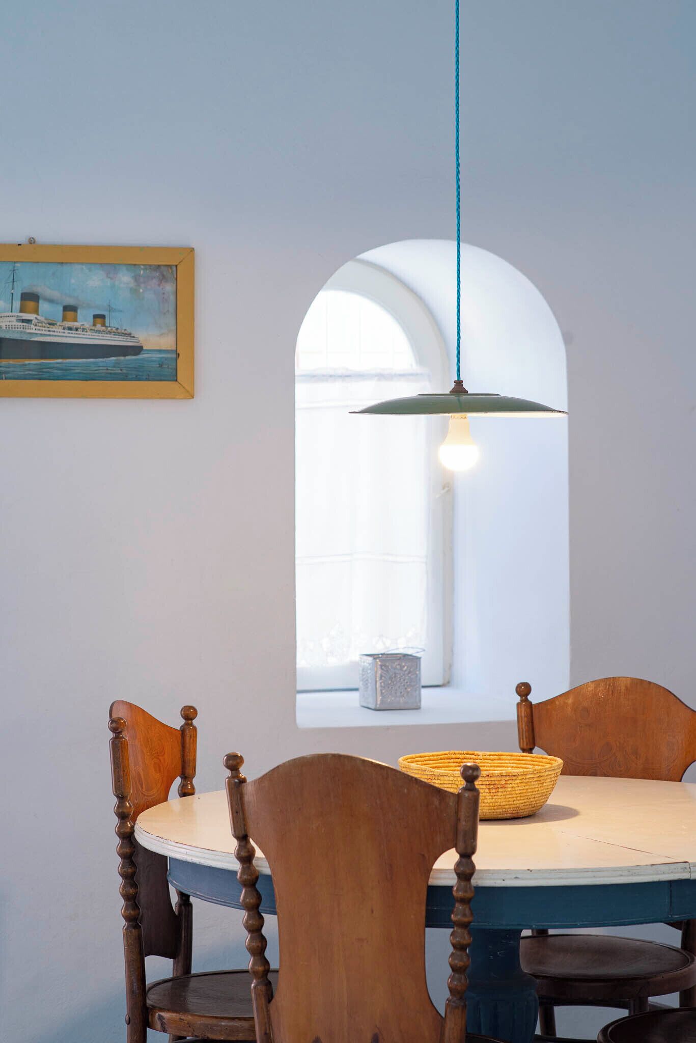 Un comedor con una mesa redonda, sillas de madera, una lámpara colgante, un ventanal con arco y un cuadro en la pared.