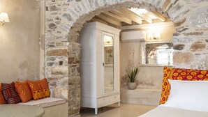 A cozy room with a stone wall, a white cabinet, a bed with colorful pillows, and a small table. - Villa Maria (Syros)
