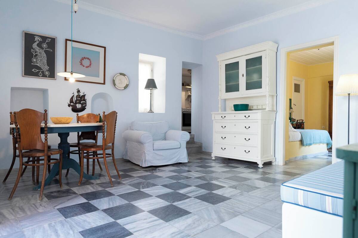 A room with a checkered floor, a dining table with chairs, a white sofa, a white cabinet, and a bed with a blue blanket.