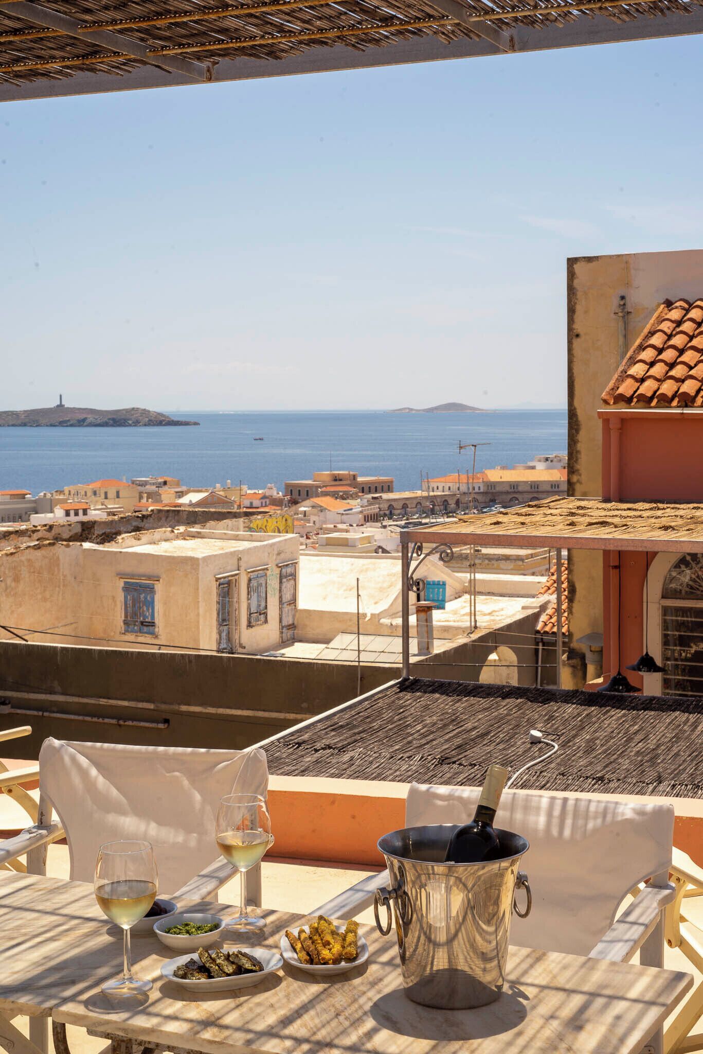 A table with a bottle of wine, two glasses, a bowl of olives, and a plate of food, overlooking a coastal town with buildings and the sea.