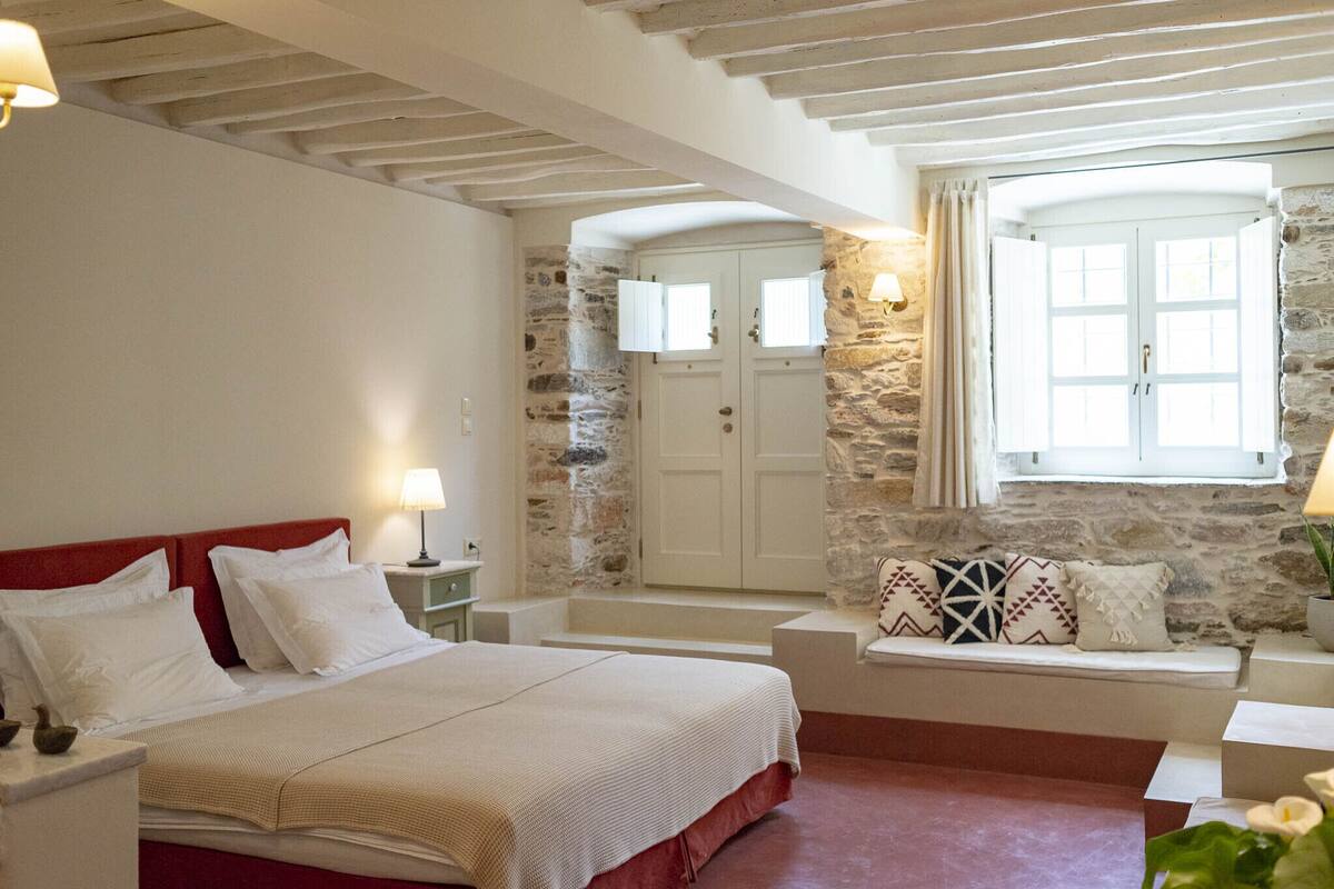 A bedroom with a bed, bedside tables, a stone wall, a window, and a bench with cushions.