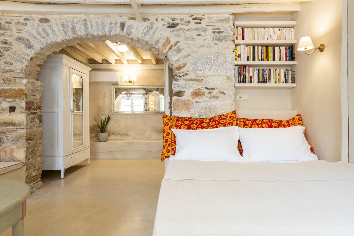 A bedroom with a stone archway, a white wardrobe, a bedside table, a bookshelf, and a window with natural light.