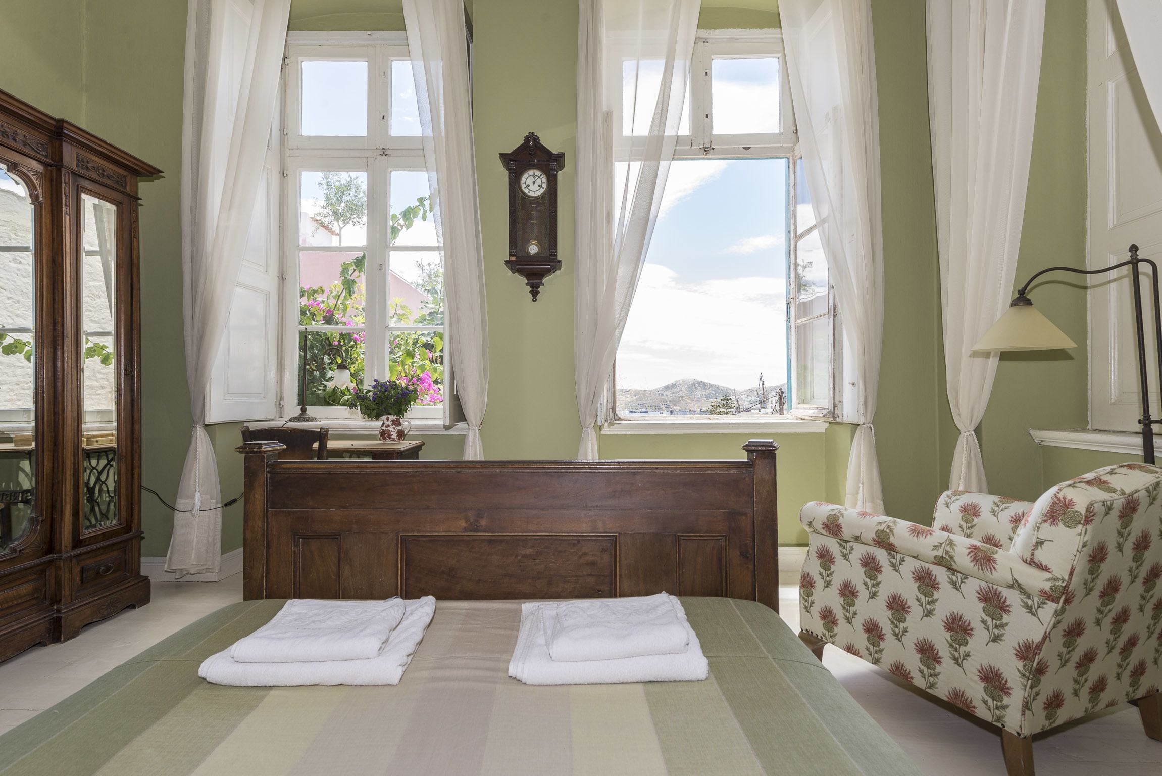 Un dormitorio con una cama de madera, dos toallas blancas, un sillón estampado y un reloj, además de un ventanal con cortinas.
