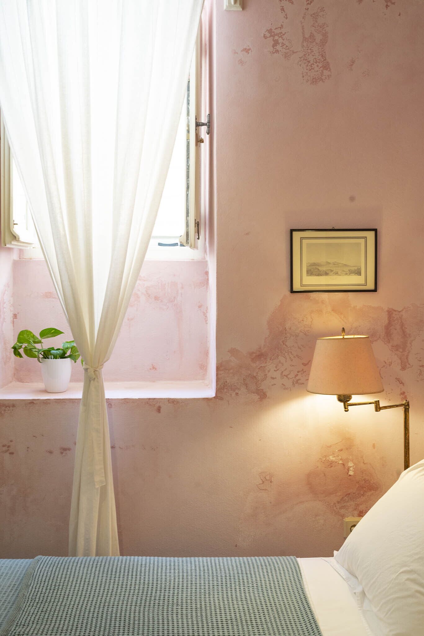 Un dormitorio con cama, lámpara de noche, ventana con cortinas e imagen colgada en la pared.