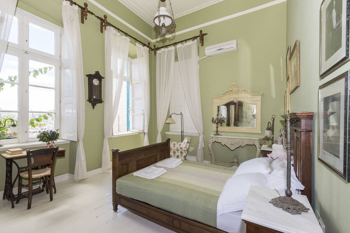 A room with a bed, a desk, and a chair. There is a window with curtains and a clock on the wall.