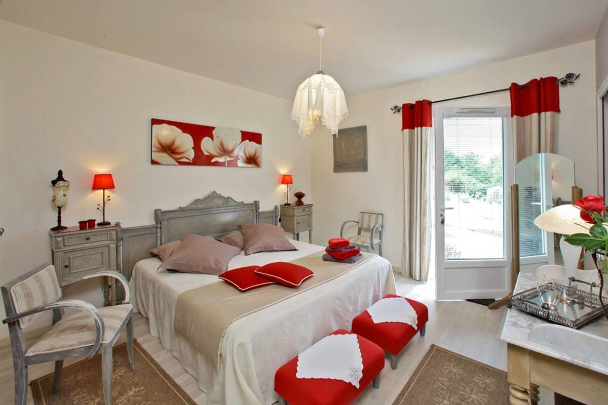 A bedroom with a double bed, two bedside tables, a chair, a desk with a mirror, a hanging light fixture, and a window with curtains.
