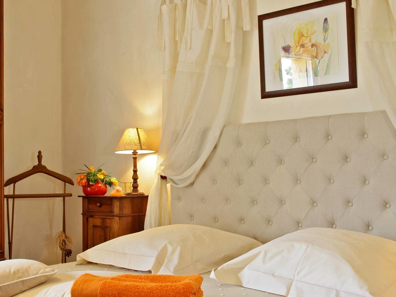 Une chambre d’hôtel avec une tête de lit capitonnée, une table de chevet en bois avec une lampe, un tableau encadré au mur et un lit avec des oreillers blancs et un plaid orange.