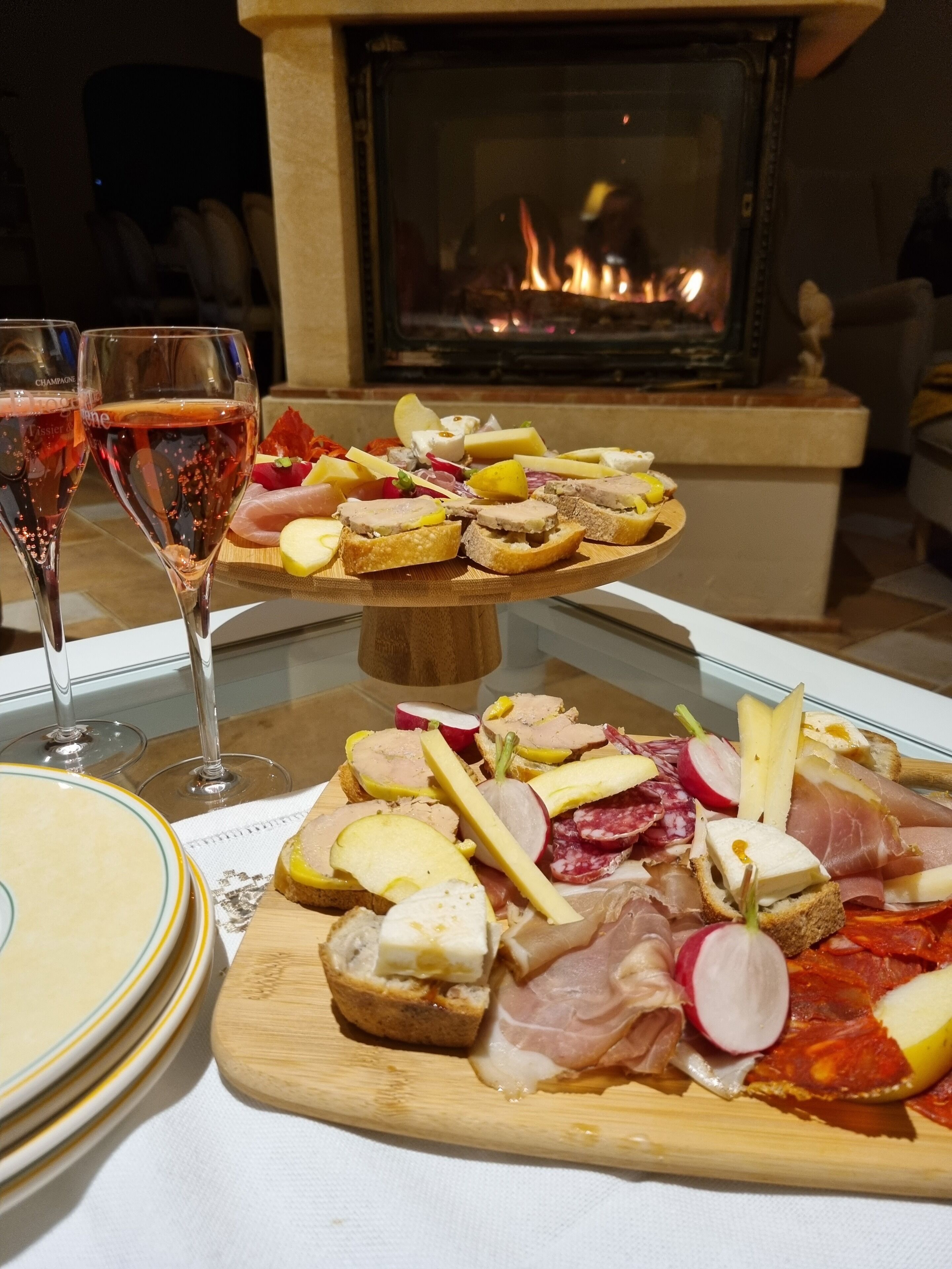 A charcuterie board with various meats and cheeses, two glasses of red wine, and a fireplace in the background.