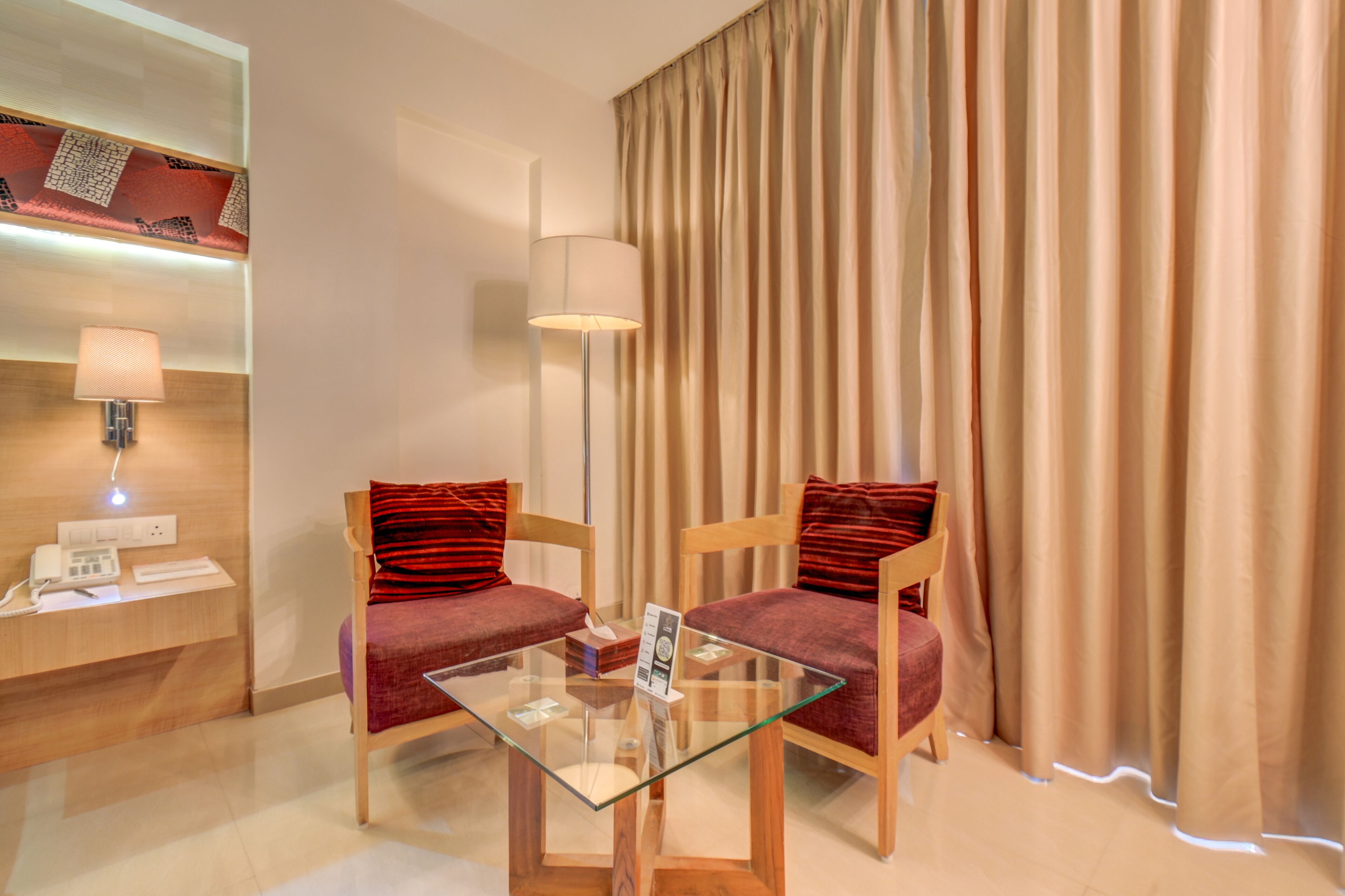 A hotel room with two wooden chairs, a glass table, a lamp, a telephone, and a curtain.