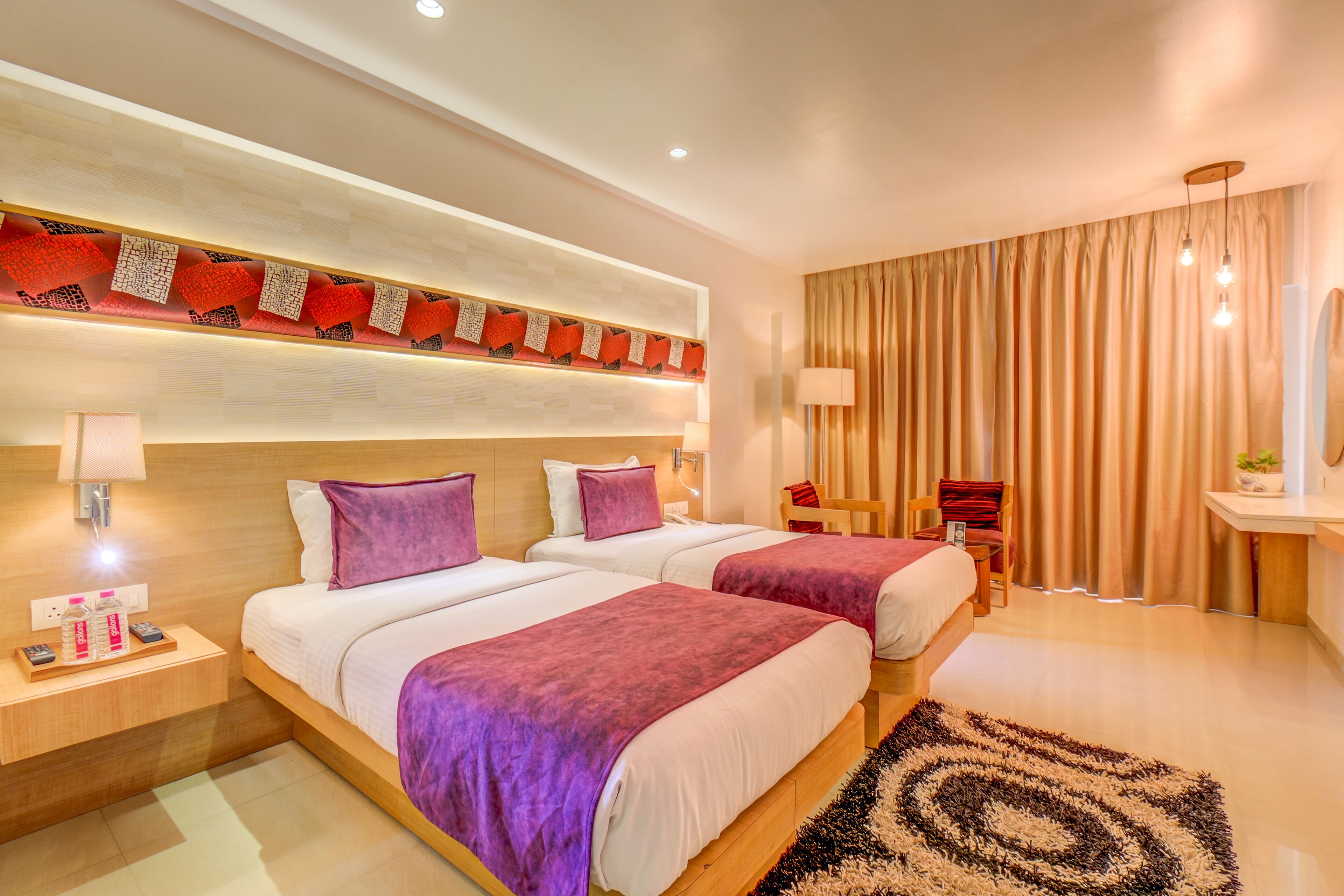 A hotel room with two beds, a wooden headboard, a nightstand with a lamp, a desk with a chair, and a large window with curtains.