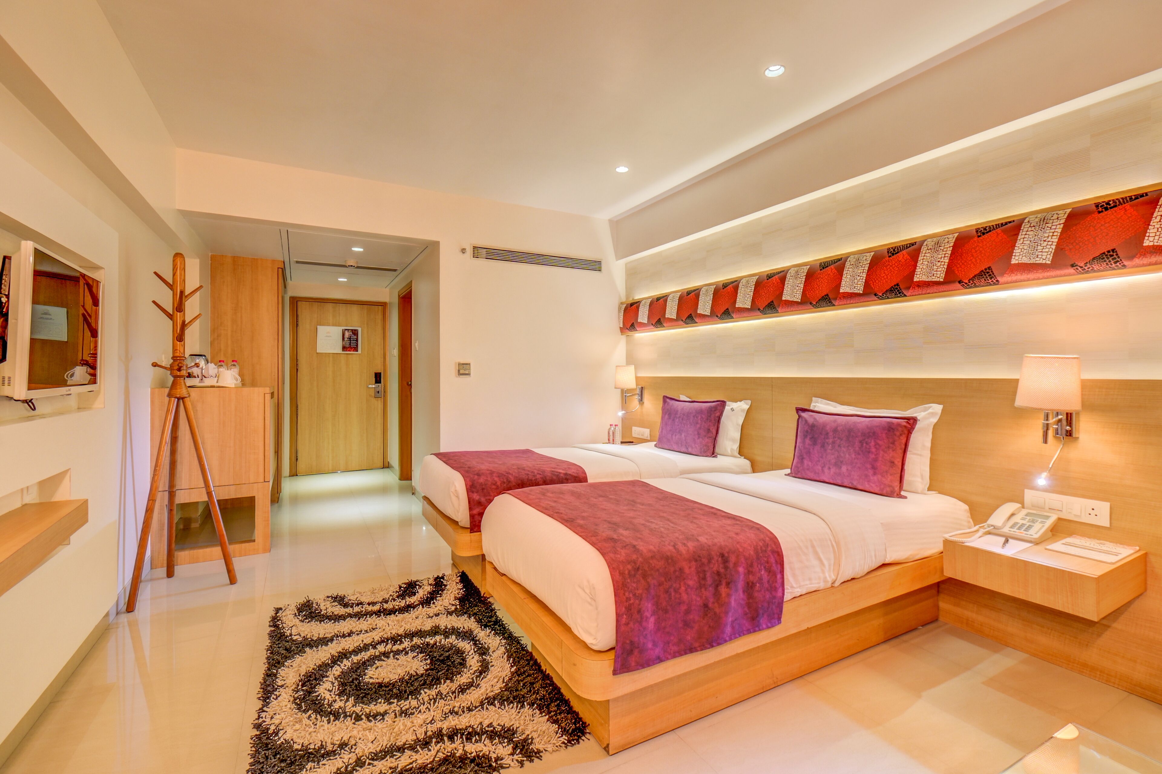 A modern hotel room with two beds, a wooden floor, a patterned rug, a wooden side table with a lamp, and a wall-mounted telephone.