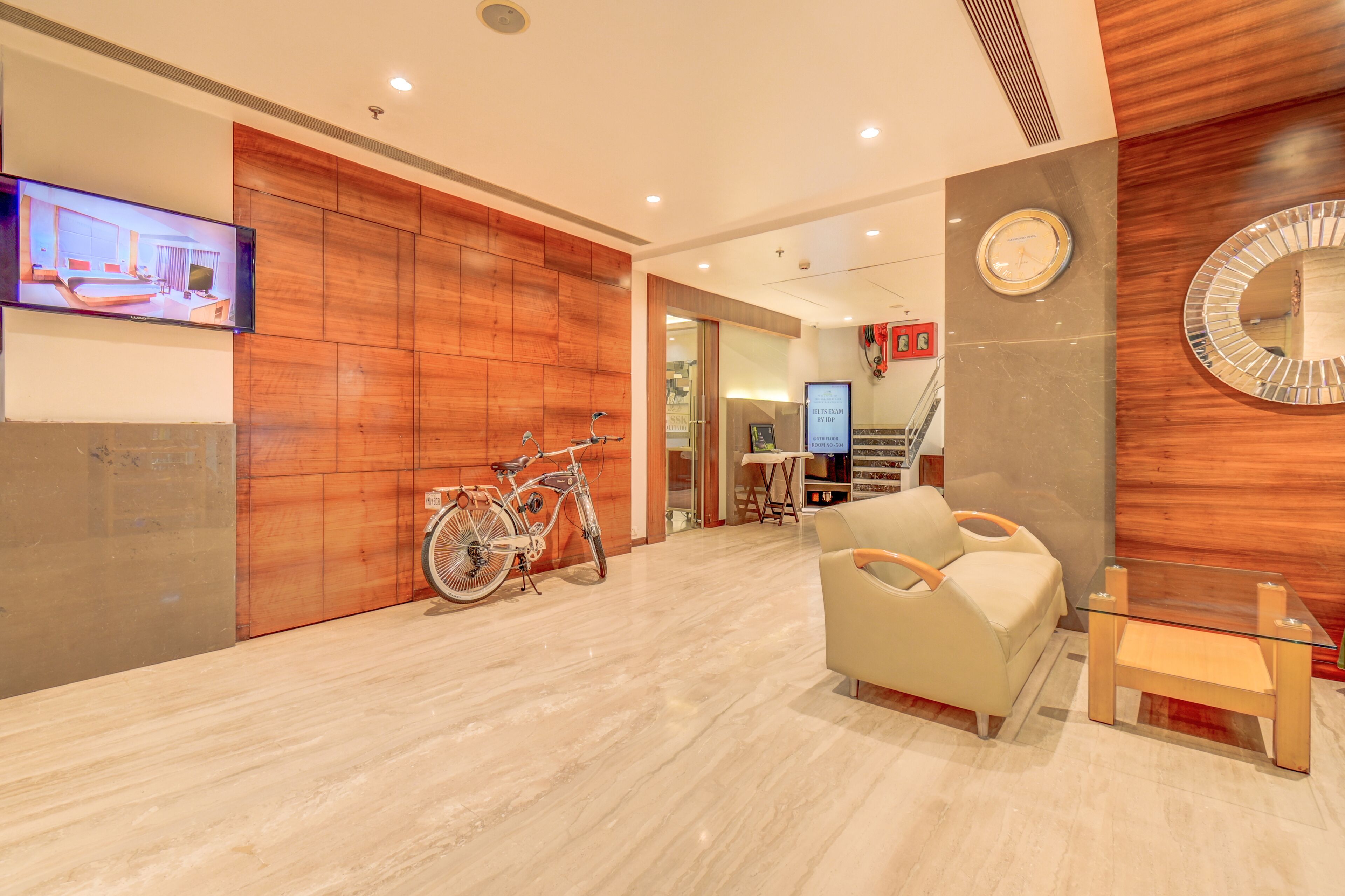 A modern hotel lobby with a wooden reception desk, a mounted television, a bicycle, a sofa, and a small table.