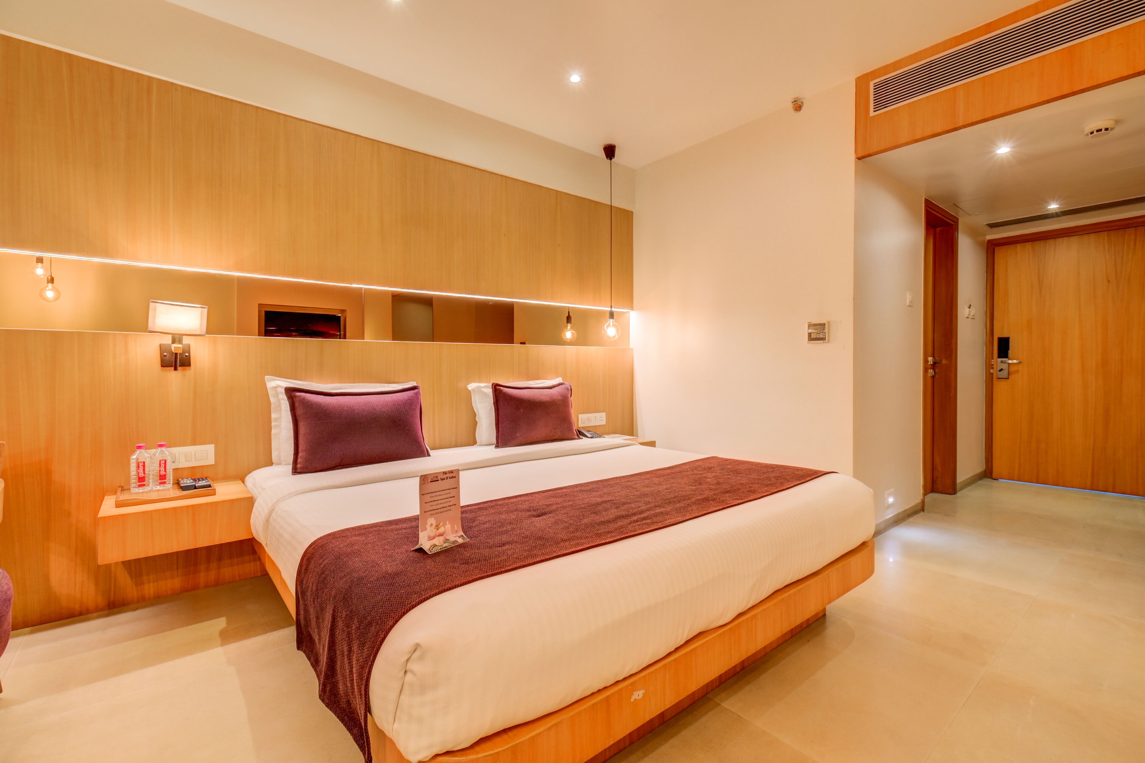 A modern hotel room with a large bed, wooden headboard, bedside tables, and a built-in TV.