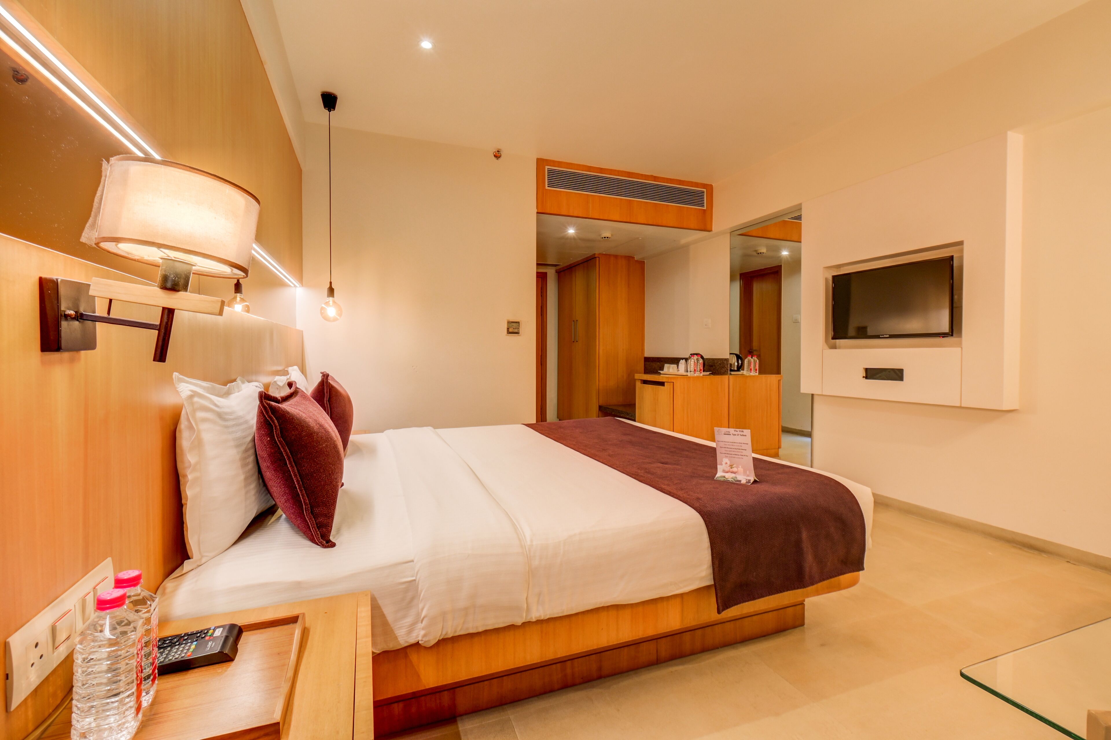 Una habitación de hotel moderna con una cama grande, una mesita de noche con control remoto, una mesa de centro de cristal, un televisor montado en la pared y un armario de madera.