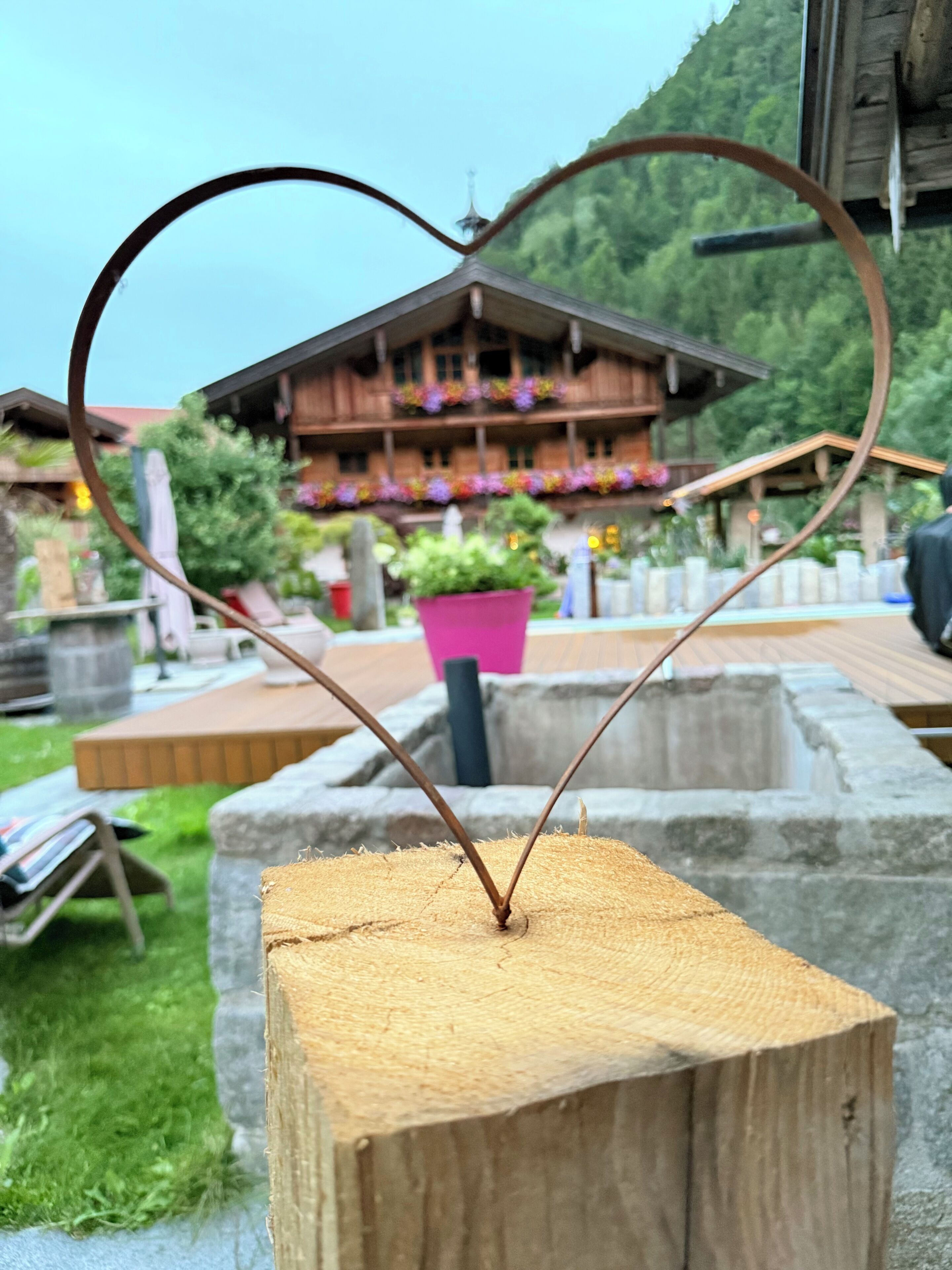A heart-shaped metal decoration on a wooden post with a wooden building and greenery in the background.