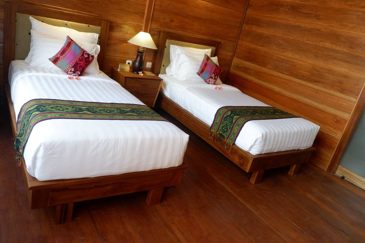 Two single beds with wooden headboards, white linens, and decorative pillows. Each bed has a bedside table with a lamp. The room has wooden flooring and walls.