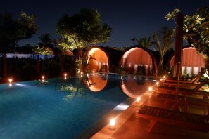 Un área de piscina nocturna con cabañas de madera y sillones de descanso.