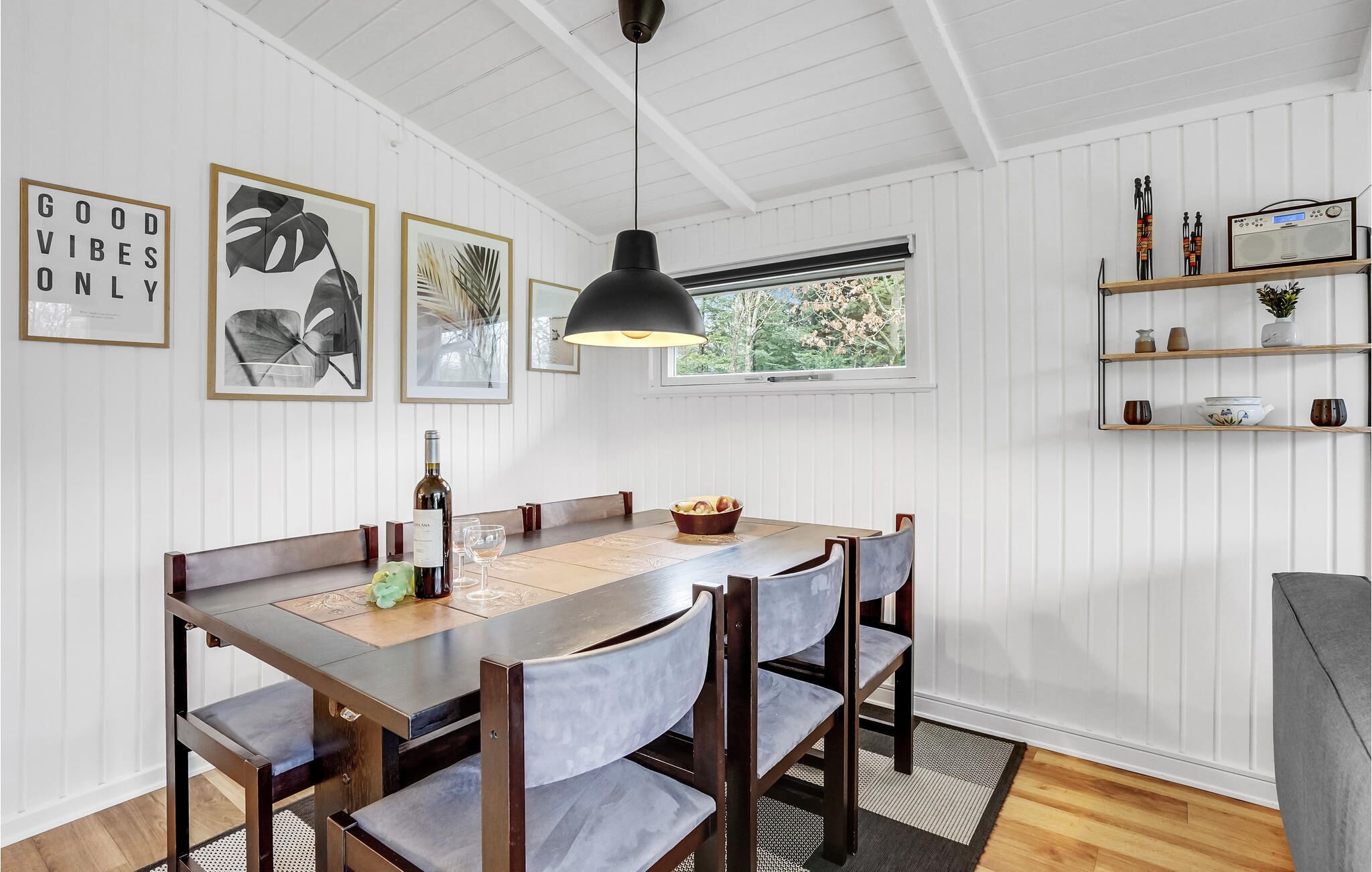 A dining area with a wooden table, chairs, and a pendant light. There are framed pictures on the wall and a window with a view of greenery.