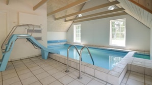 Una piscina con uno scivolo blu e due scalette, pavimento piastrellato e ampie finestre.