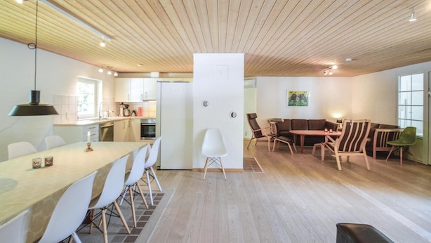 Un interno moderno con soffitto in legno, una zona pranzo con sedie bianche, una cucina con mobili bianchi e un angolo salotto con diverse sedie.