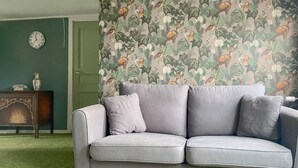 Un canapé gris avec des coussins, dans une pièce dotée d’une moquette verte et d’un papier peint à motifs floraux.