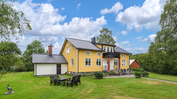 Une maison jaune à deux étages, dotée d’un balcon, d’un petit abri de jardin et d’une pelouse aménagée avec du mobilier d’extérieur.