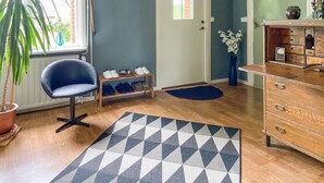 Une pièce avec une chaise bleue, un porte-chaussures en bois, un tapis à motifs, une commode en bois et une plante en pot.