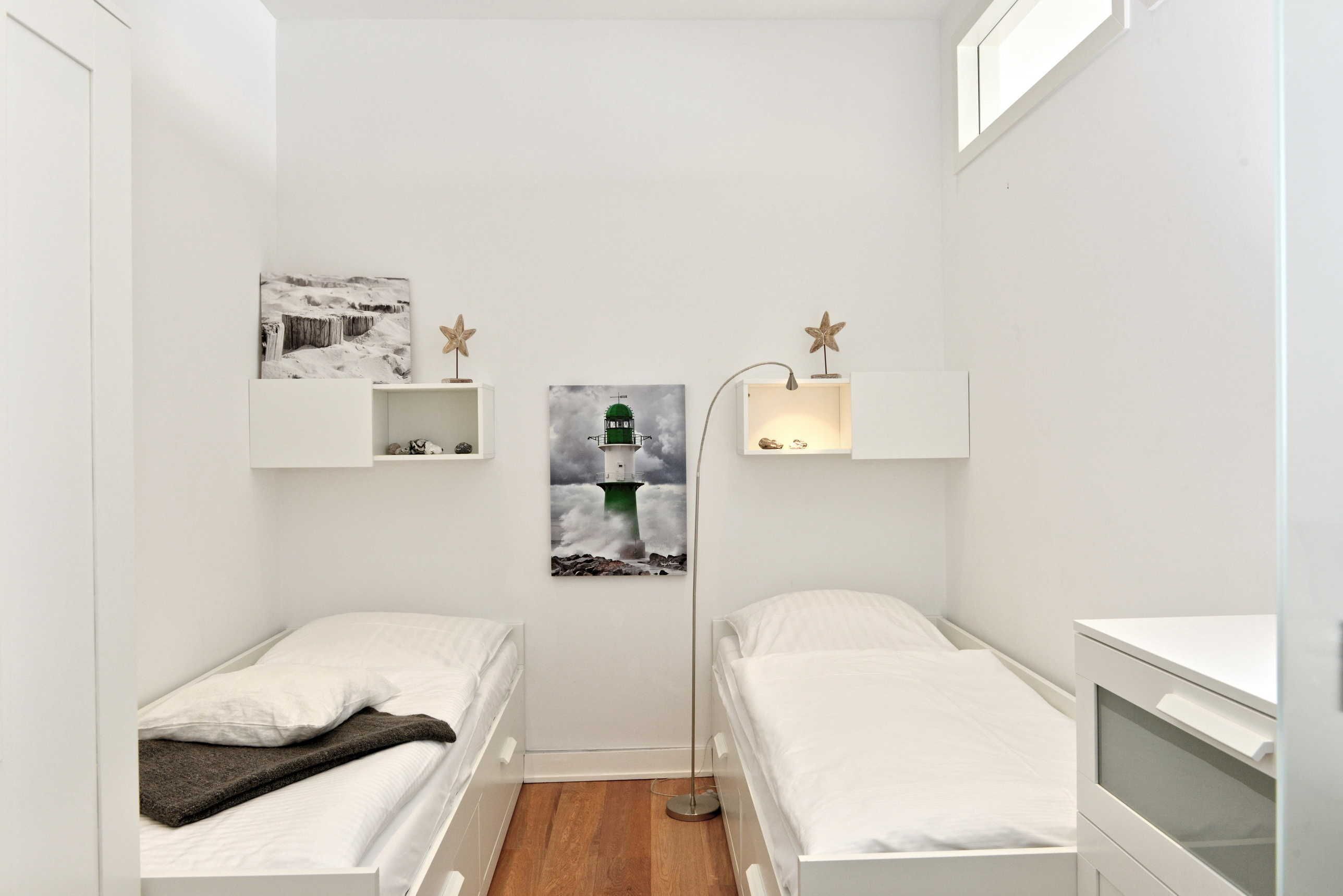 Una habitación pequeña y blanca, con dos camas, una lámpara de pie y estantes con fotografías.