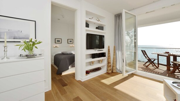 Un salón moderno con televisor, estanterías blancas, una planta y un balcón con vistas.