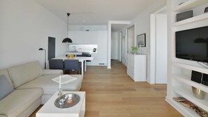 Una sala moderna con sofá, mesa de centro y mueble para la televisión. Se observa un área de cocina con mesa y sillas al fondo.