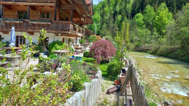 A wooden chalet with a balcony overlooking a river, surrounded by lush greenery and a mountainous backdrop.