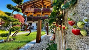 A wooden chalet with a balcony, surrounded by a well-maintained garden and a stone wall with strawberries.