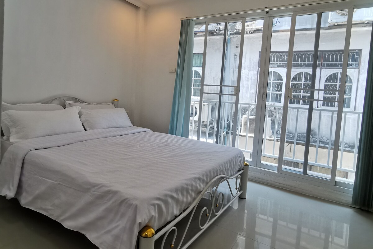 Ein Schlafzimmer mit Bett, Deckenventilator, Fenster mit Vorhängen und Blick ins Freie.