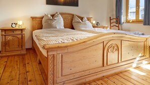 Ein hölzernes Bett mit karierter Bettdecke, zwei Nachttische mit Lampen, ein Beistelltisch, ein Holzstuhl und ein Fenster mit Vorhängen.