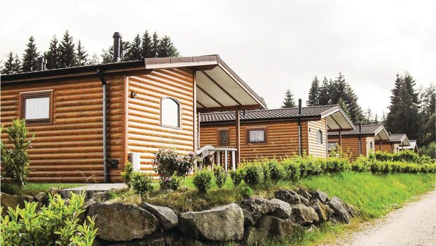 A row of log cabins with a stone wall in front, a gravel path to the right, and a grassy area with bushes.