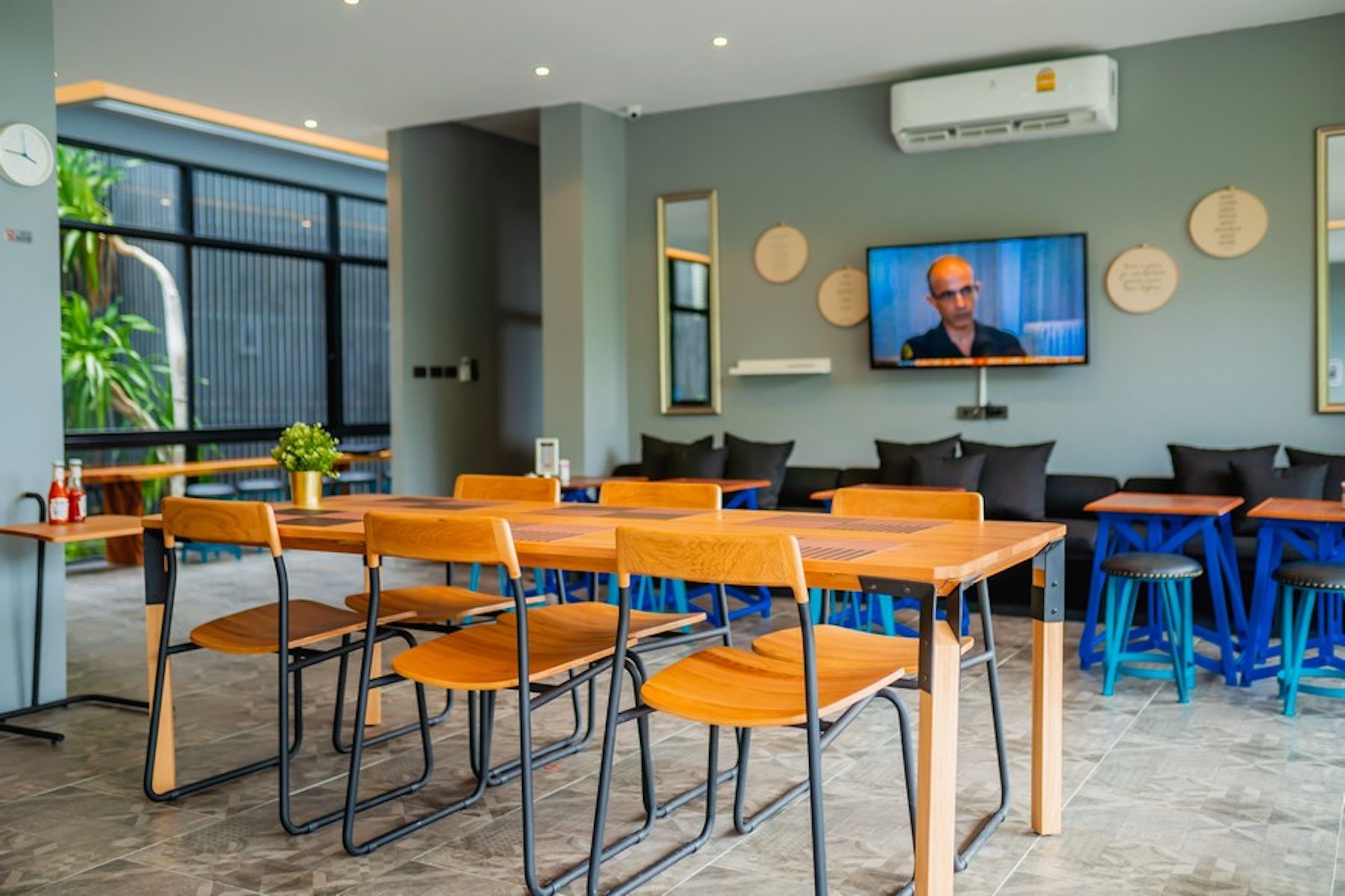 A modern dining area with wooden tables and chairs, a television screen, and a large window with plants.