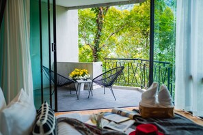 Un balcon avec une table et des chaises, une vue sur les arbres, et un lit avec un livre et un chapeau.