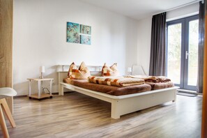 A modern bedroom with a wooden floor, a white bed, and a view of the outdoors through a large window.