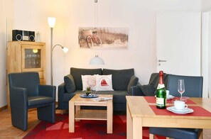 Ein Wohnzimmer mit einer Couch, zwei Sesseln, einem Couchtisch, einem Esstisch mit einer Champagnerflasche und einem Glas sowie einer Lampe.