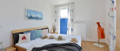 Un dormitorio ordenado con cama, mesitas de noche, una ventana con cortinas, un cuadro en la pared y un perchero.