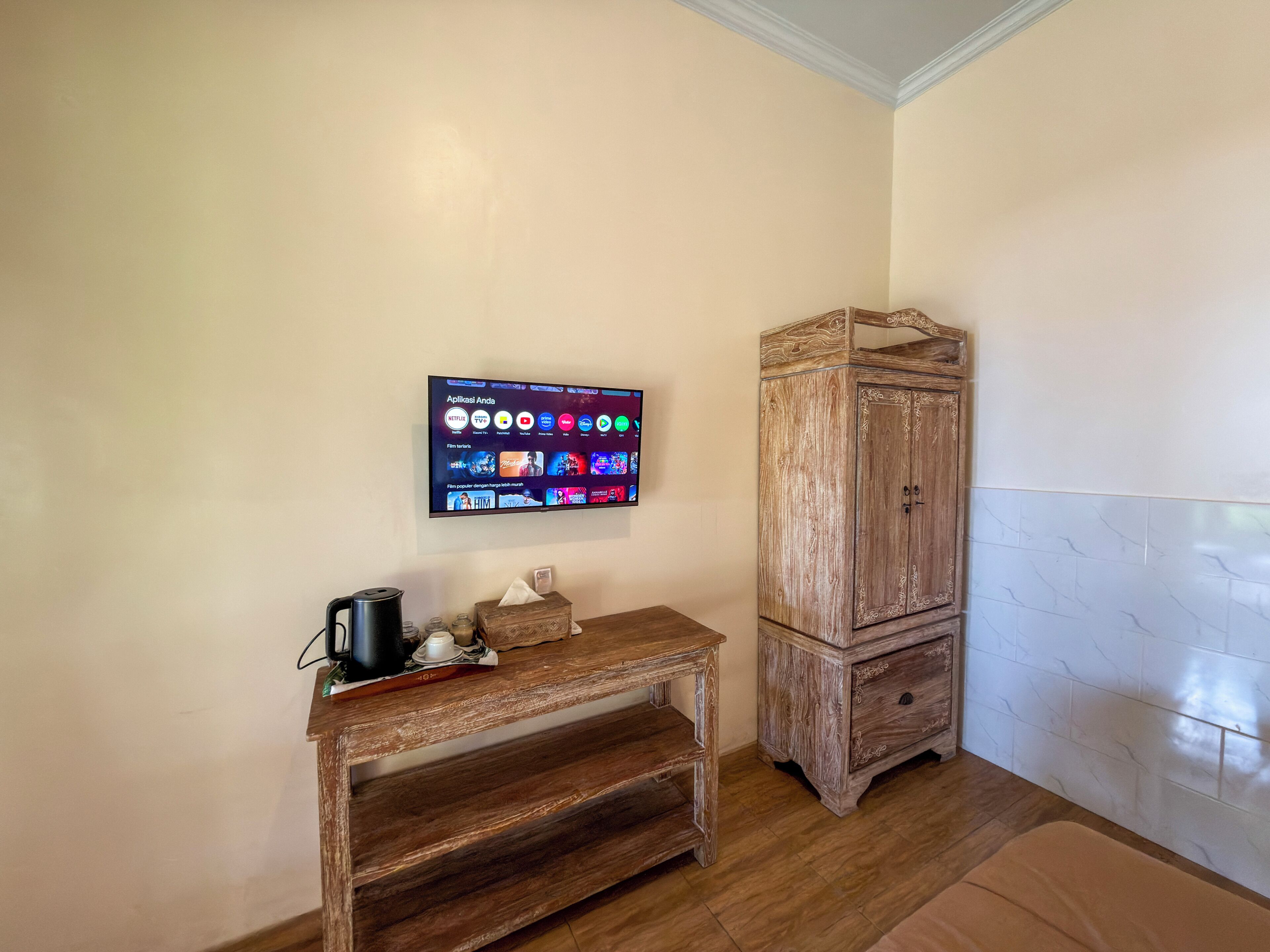 A room with a wooden cabinet, a small wooden table with a kettle and a tissue box, and a flat-screen TV mounted on the wall displaying various apps.