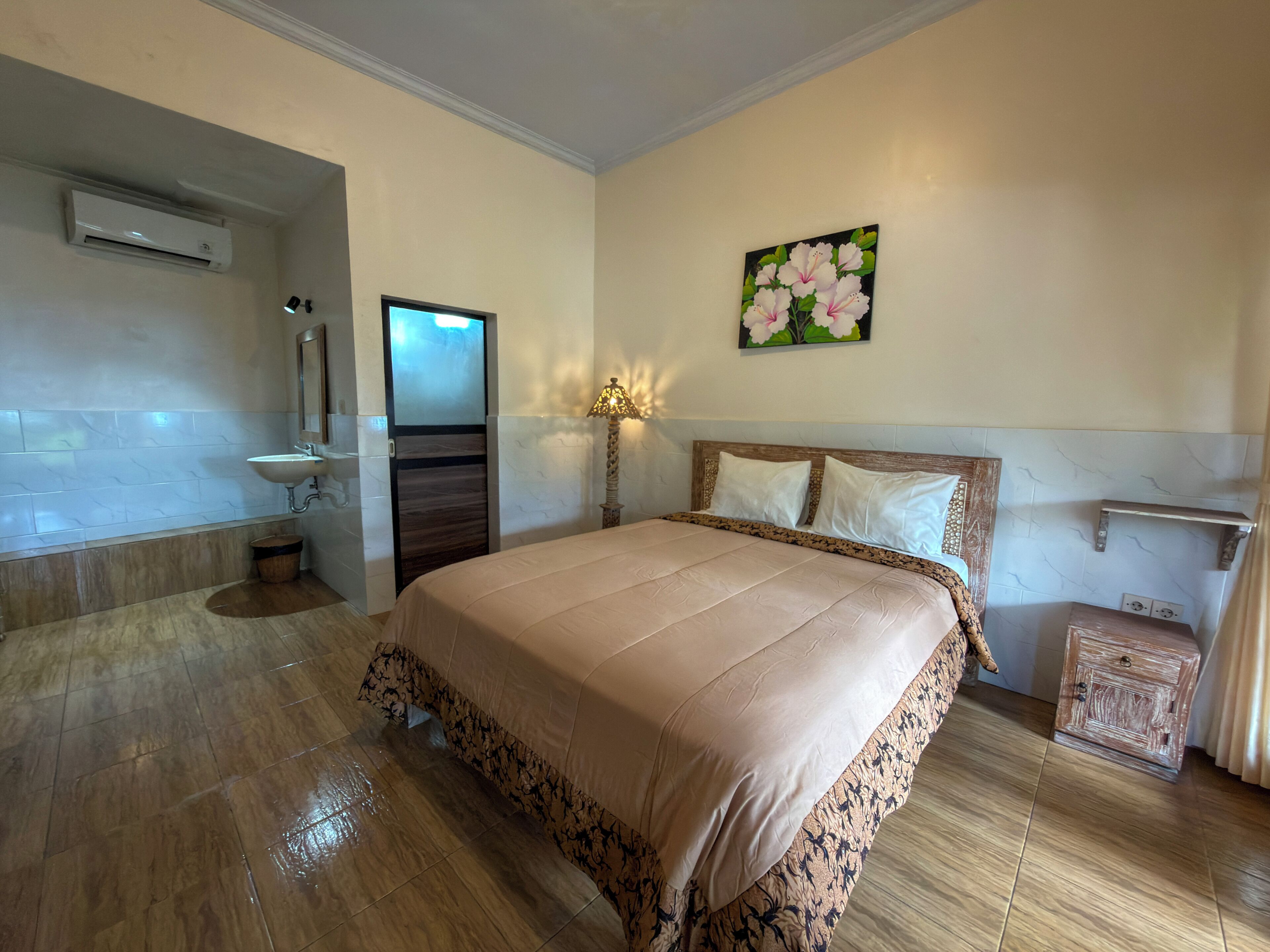 A bedroom with a bed, a nightstand, a lamp, a painting, a bathroom with a sink, and an air conditioning unit.