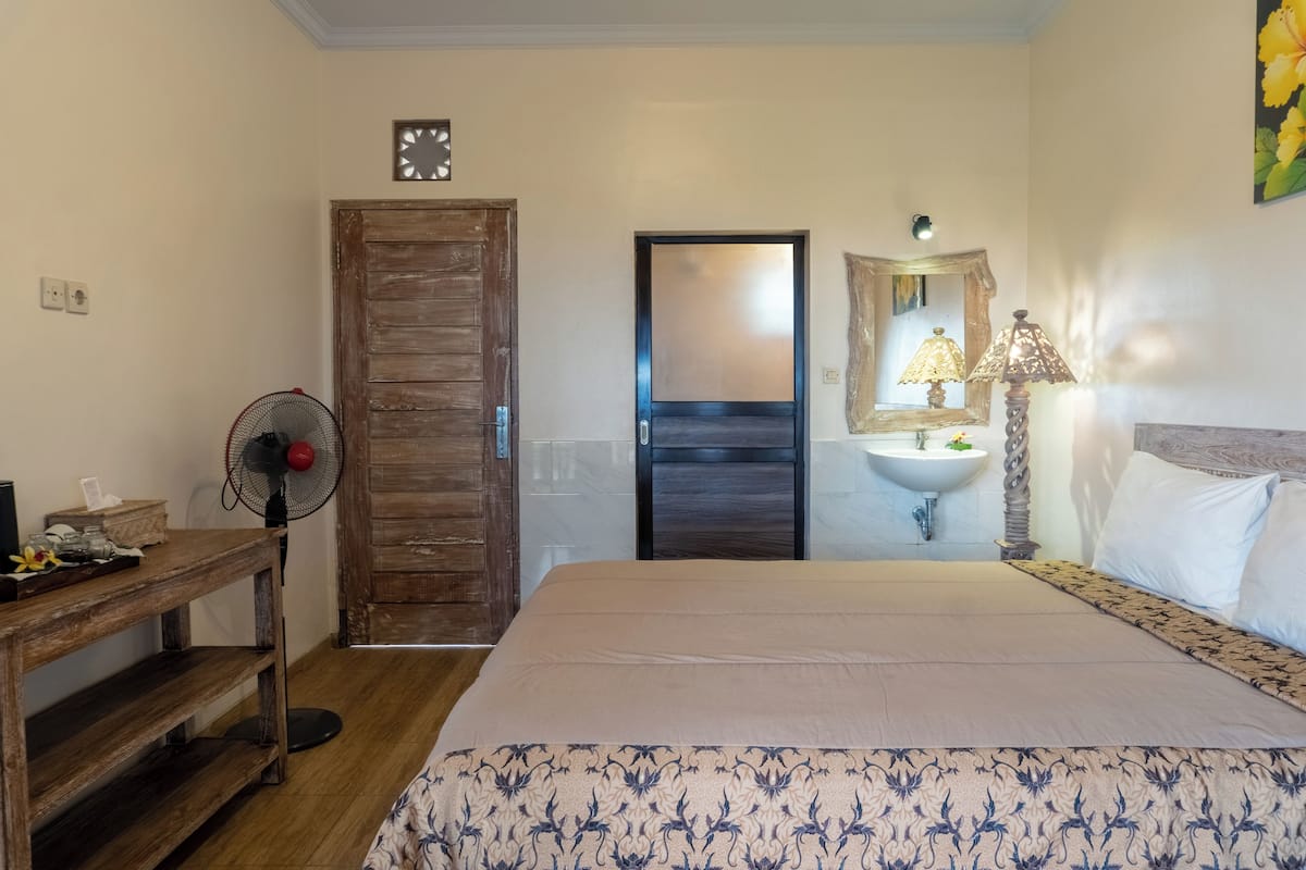 A bedroom with a bed, bedside table, wooden door, and a small table with a fan.