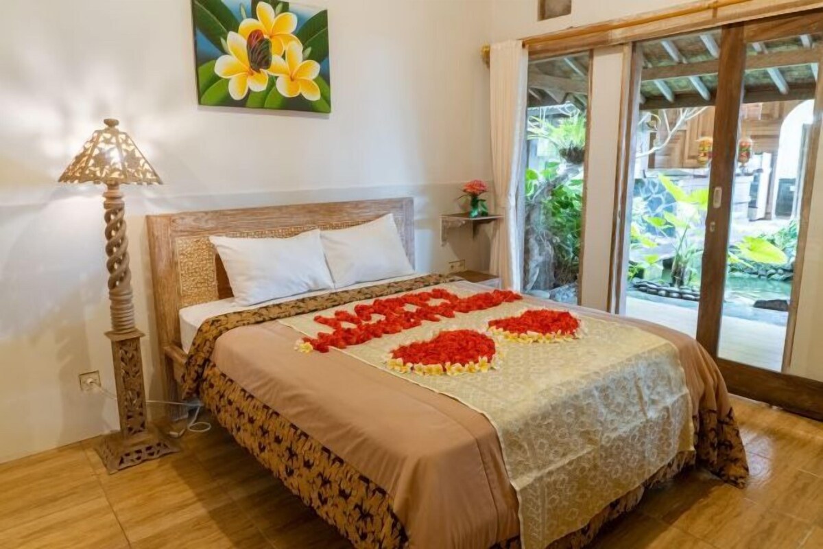 A hotel room with a bed, a nightstand, a lamp, a painting of flowers, and a view of the outdoors.